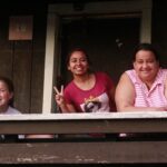 Three people smiling and posing from a balcony, with one making a peace sign. The setting appears to be a rustic wooden building.
