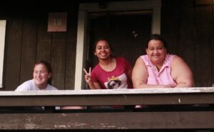 Three people smiling and posing from a balcony, with one making a peace sign. The setting appears to be a rustic wooden building.