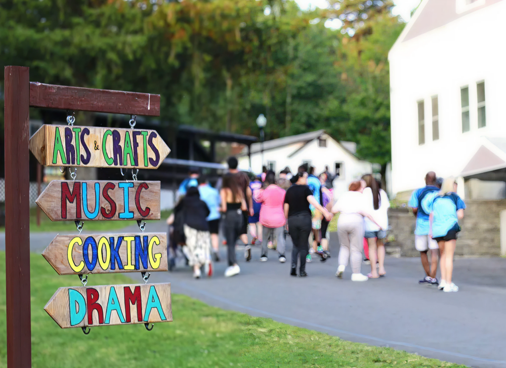 A sign at Camp Loyaltown that reads "Arts & Crafts" "Music" "Cooking" and "Drama" with arrows.