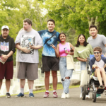 A diverse group of seven smiling individuals, including one person using a wheelchair, pose together outdoors on a paved path surrounded by trees.