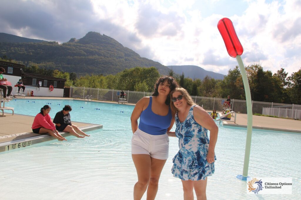 IMG_7858 | Citizens Options Unlimited Two women smile together while standing in the shallow water of an outdoor swimming pool, with a splash pad feature, onlookers, and a scenic mountain landscape in the background.