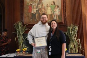 Man in a gray hoodie holds a certificate while posing with a smiling woman at an awards ceremony.