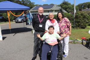 Four adults posing for a photo at an outdoor event; a person in a wheelchair sits front center, others stand behind under a blue tent with decorations.