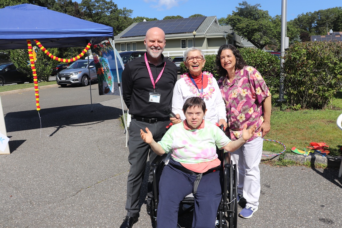 Four adults posing for a photo at an outdoor event; a person in a wheelchair sits front center, others stand behind under a blue tent with decorations.