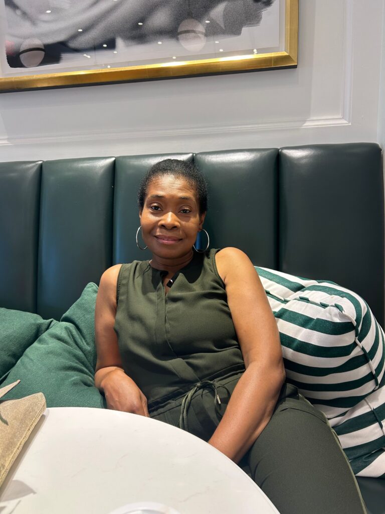Elizabeth Delisca | Citizens Options Unlimited Woman with short hair and hoop earrings relaxing on a green leather bench in a cafe, striped pillow behind her.