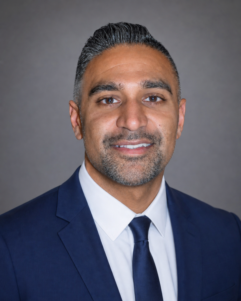 Ishri Prashad | Citizens Options Unlimited Professional head-and-shoulders portrait of a smiling man in a navy suit and white shirt against a gray backdrop.