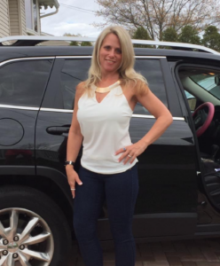 Woman posing by a black SUV with the passenger door open, wearing a white sleeveless top and dark jeans on a driveway.