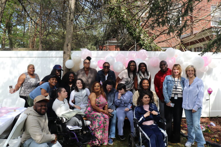 Diverse group posing for a celebration outdoors with pink and white balloons and a white fence behind them.