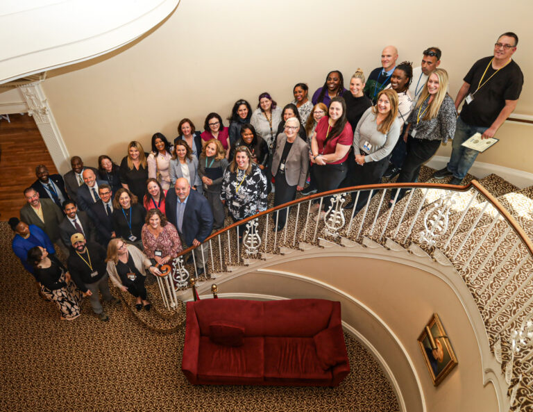 Group of diverse professionals posing on a grand curved staircase, looking up at the camera, wearing lanyards and badges.