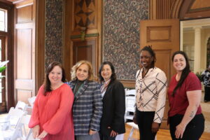 Five women stand side by side posing for a group photo in a historic room with wood paneling and decorative wallpaper, all smiling at the camera.
