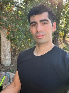 Portrait of a man in a black t-shirt standing outdoors with trees and foliage in the background. A dog on a leash is partially visible at the lower left.