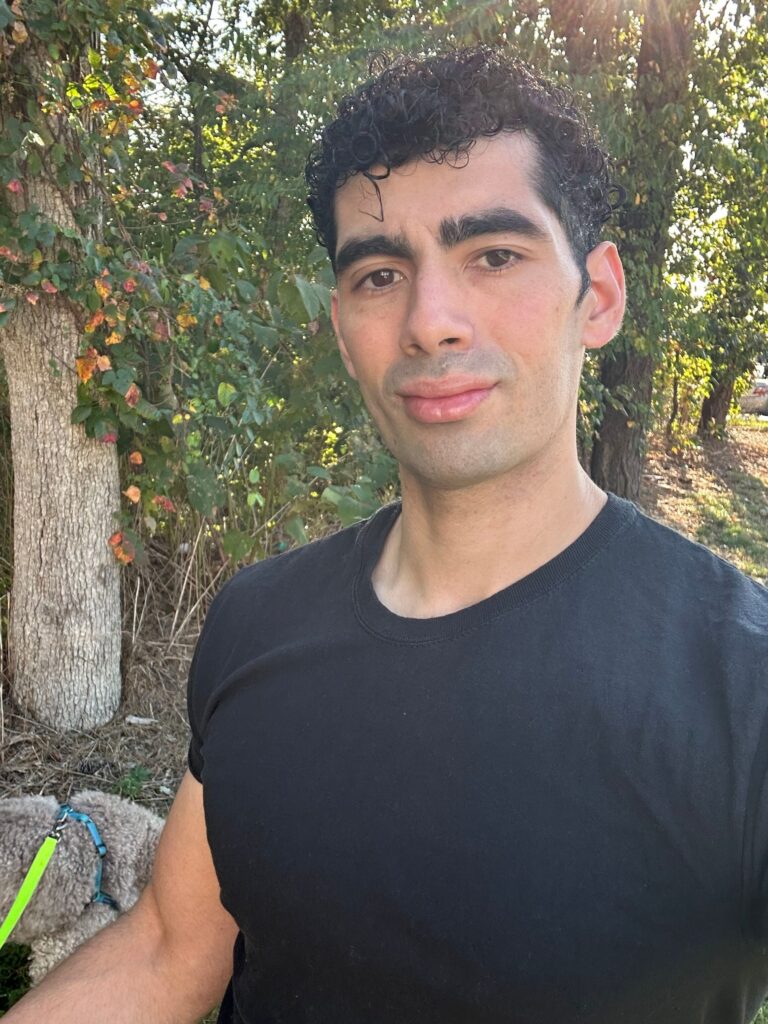 Portrait of a man in a black t-shirt standing outdoors with trees and foliage in the background. A dog on a leash is partially visible at the lower left.