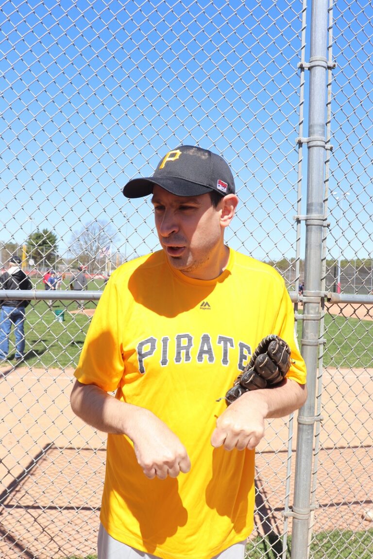 Man wearing a yellow Pirates T-shirt and black cap stands by a chain-link fence, holding a baseball glove.