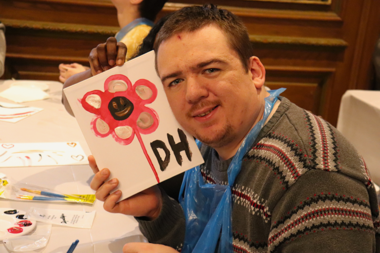 Man with short hair and light facial hair smiles while holding a white canvas depicting a pink flower with a brown center at a craft table.