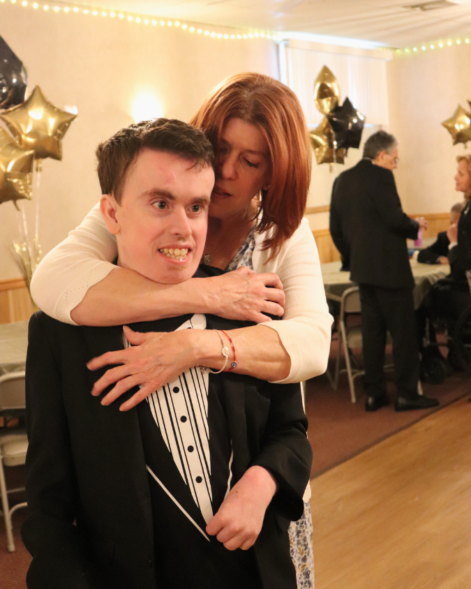 Woman in a white top hugs a young man in a black tuxedo from behind at a celebration, with gold star balloons in the background.