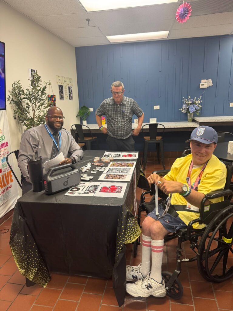 Three men at a table with event materials; a man in a wheelchair wearing a yellow jacket sits on the right while two others sit/stand nearby, in a room with a blue decorative wall.