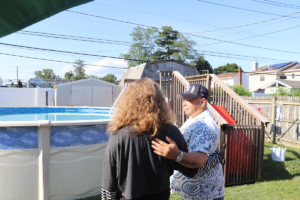 Man wearing a patterned shirt and cap hugs a woman by an above-ground pool in a sunny backyard, with a wooden deck nearby.