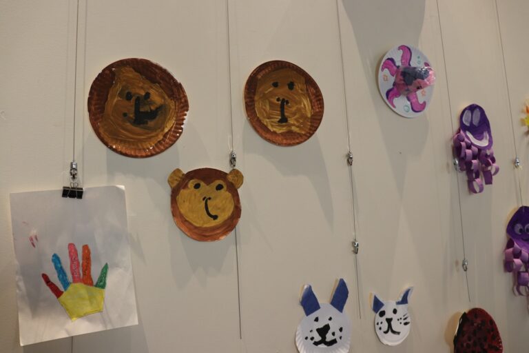 Children's art display on lockers: painted paper-plate bear faces and animal crafts hanging from strings beside a handprint drawing.