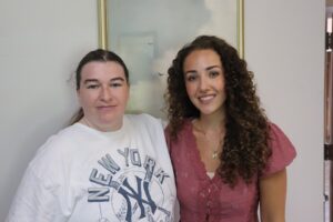 Two women posing together indoors, one in a white graphic tee and the other in a pink blouse, both smiling at the camera.