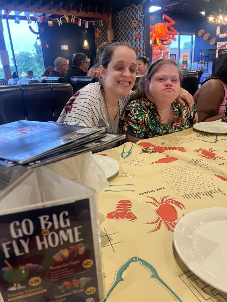 Two women sit close together at a seafood restaurant table, smiling playfully for the camera with one grinning widely and the other puffing her cheeks, creating a goofy moment in the foreground.