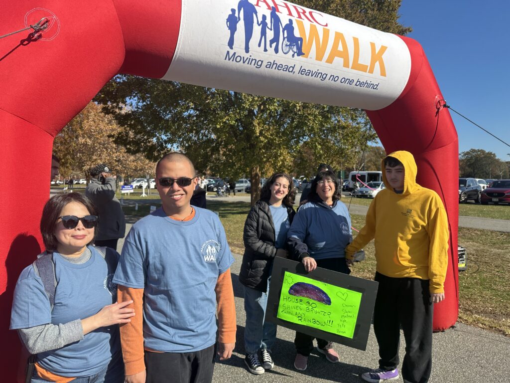 Group of volunteers posing under a red inflatable arch at an outdoor charity walk, wearing blue shirts; a bright green poster with a rainbow drawing is visible.