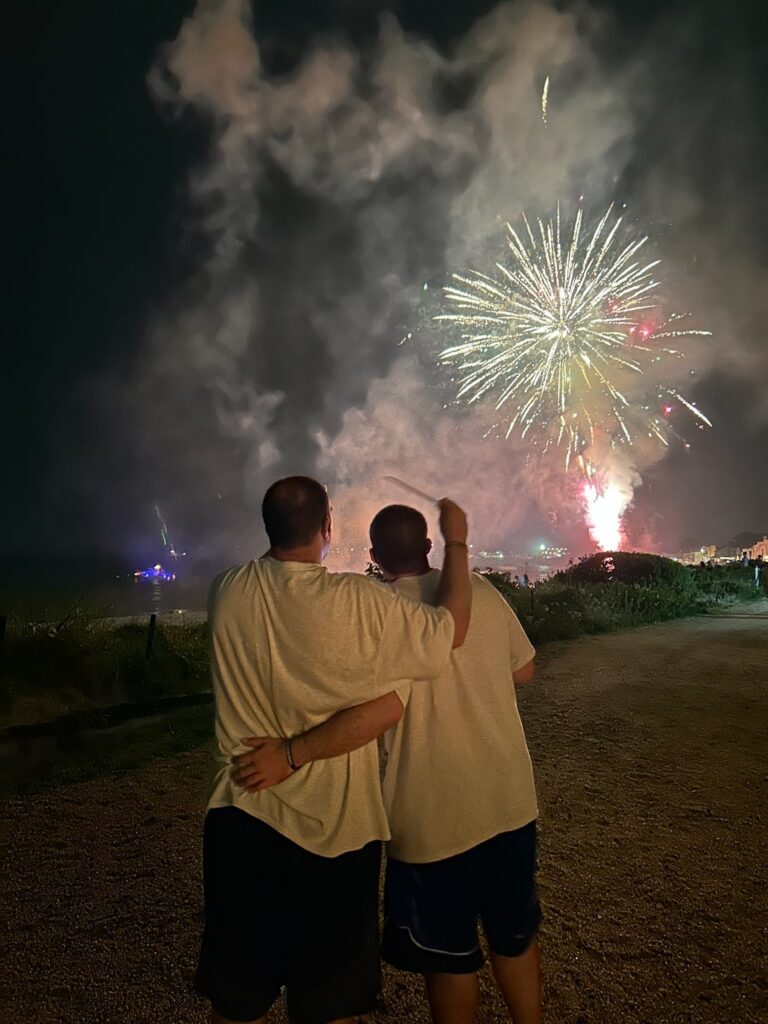 Two people stand with arms around each other, watching a fireworks display at night on a beach/park.