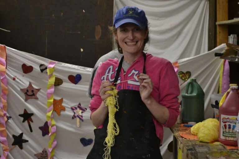 Smiling woman in a pink hoodie and black apron holds yellow yarn strands in a craft studio with colorful decorations behind her.