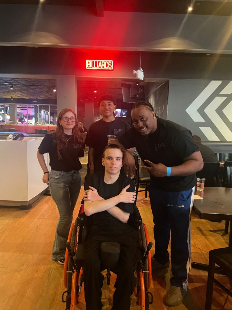 Four friends posing together indoors at a recreation venue, with one person in a wheelchair at the front and a red neon 'BILLIARDS' sign in the background.