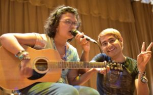 A camper plays guitar and sings while a counselor holds the microphoner
