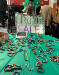 Ruth Henderson holding a sign that says "Ruth's Sale" at a table where she has her bracelets on display.