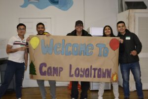 Camp Loyaltown staff members hold up a hand painted sign reading "WELCOME TO CAMP LOYALTOWN"